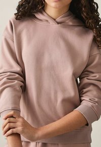 En person med krøllet hår bærer en pink hættetrøje, hænderne sammenfoldet ved albuen, mod en neutral baggrund. Fokus er på detaljerne på hættetrøjen.