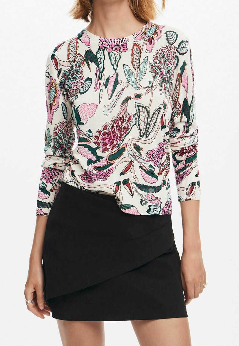 Femme portant un haut blanc à manches longues avec un imprimé floral rose et vert et une mini-jupe asymétrique noire sur fond blanc.