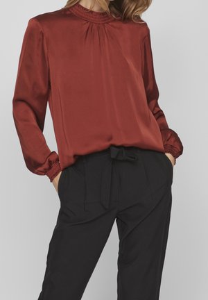 Kvinde iført en rustrød satinbluse med rynkede manchetter og halsudskæring, kombineret med højtaljede sorte bukser bundet i taljen.