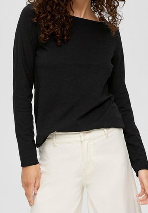 Vrouw draagt een zwart top met lange mouwen en een wit broek met hoge taille, staand tegen een effen lichte achtergrond.