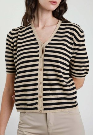 Donna che indossa un cardigan beige e nero a righe orizzontali con maniche corte, abbottonato, con pantaloni beige, braccia posizionate dietro la schiena.