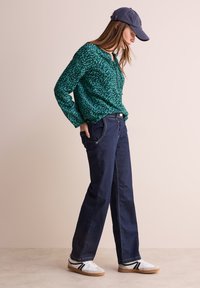Blusa verde a fantasia, jeans a gamba larga blu scuro, sneakers bianchi con strisce nere e cappellino blu navy. Tessuto testurizzato ed elementi di design casual.