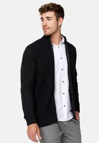 Gilet noir tricoté avec un devant texturé, des poignets côtelés et une fermeture éclair. Porté par-dessus une chemise blanche avec des boutons marron, complété par un pantalon gris.