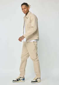 Homme marchant, portant une veste et un pantalon beiges, une chemise blanche, et des baskets noir, blanc et bleu, sur un fond gris clair uni.