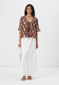 Blusa marrone a fantasia con maniche corte e un nodo frontale, abbinata a una lunga gonna bianca in pizzo. La modella tiene una borsa color crema e indossa sandali.