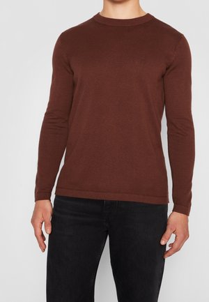 Mand iført en tætsiddende brun langærmet sweater og sorte jeans, stående mod en ensartet lys baggrund.