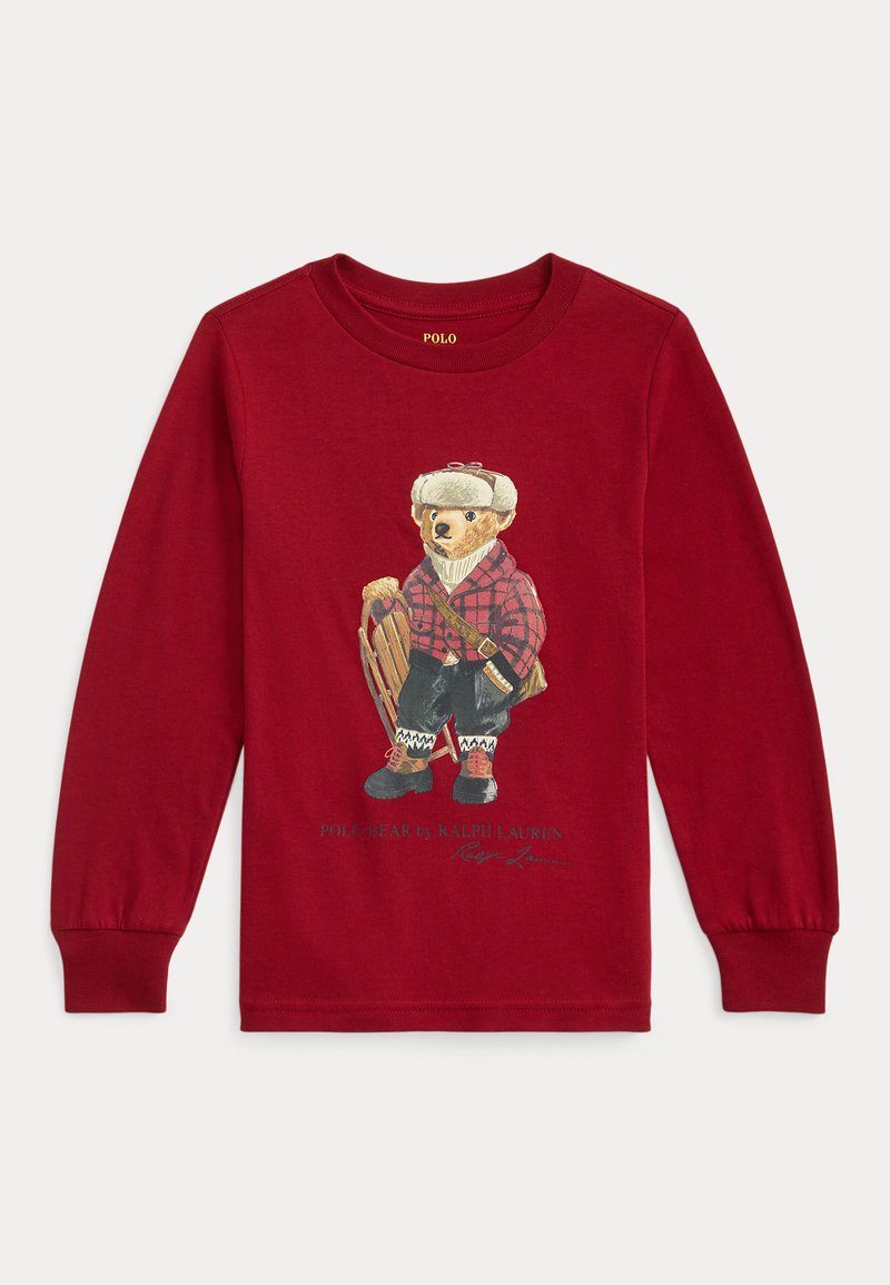 Piros hosszú ujjú póló, amelyen egy kockás inges medve grafikája látható, aki egy szánkót tart, fekete és piros díszítésekkel, alatta fehér szöveggel.