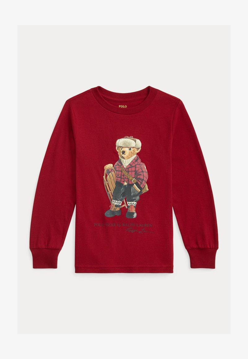 Piros hosszú ujjú póló, amelyen egy kockás inges medve grafikája látható, aki egy szánkót tart, fekete és piros díszítésekkel, alatta fehér szöveggel.