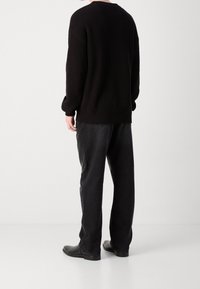 Schwarzer Rippstrickpullover mit fallenden Schultern, kombiniert mit locker sitzenden schwarzen Hosen und schwarzen Schuhen, von hinten vor einem weißen Hintergrund betrachtet.