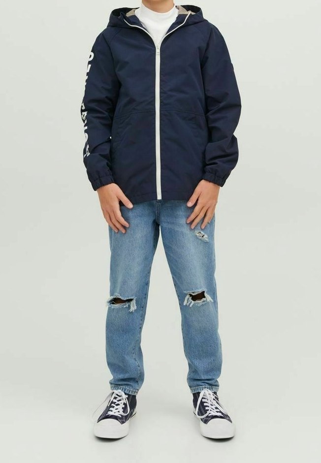 Osoba v tmavě modré bundě s kapucí a bílým zipem, bílém roláku, roztrhaných světle modrých džínách a černo-bílých teniskách, stojící čelem vpřed.