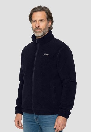 Giacca in pile blu navy con collo alto e chiusura con zip, caratterizzata da una texture morbida e un piccolo logo sul petto. Indossata con jeans azzurri.