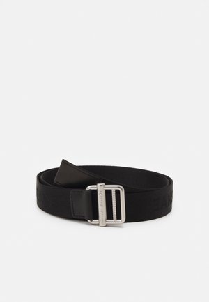 Ceinture en tissu noir avec boucle rectangulaire en métal gravée de "Tommy Jeans", enroulée sur un fond blanc.