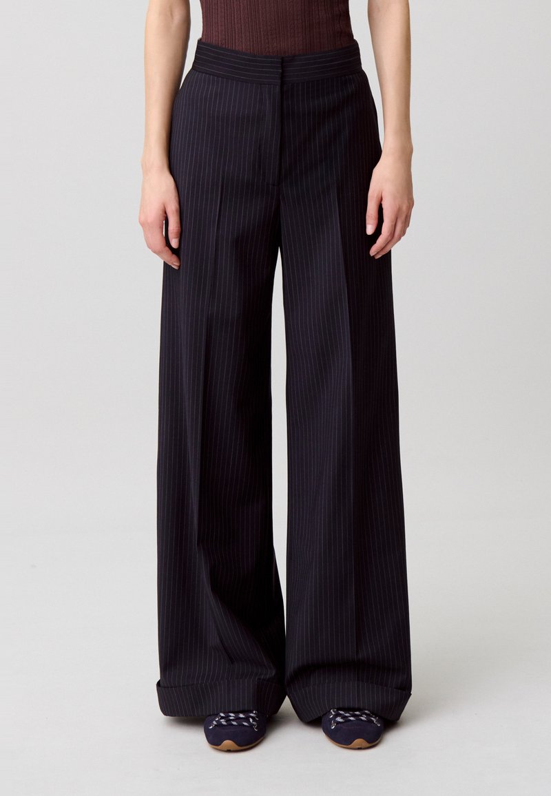 Pantaloni a gamba larga in tessuto blu navy scuro con righe verticali sottili. Presentano una vita alta e una texture liscia, abbinati a scarpe tonde blu navy.
