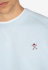 Jasnoniebieski sweter z okrągłym dekoltem i ściągaczami. Posiada małe, czerwone haftowane logo skrzyżowanych kijów golfowych i czapki.