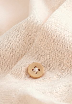 Béžová látka s našitým světle hnědým kulatým knoflíkem, zobrazující podrobnou tkanou texturu a šití.