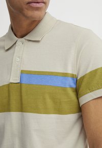 Polo skjorte i lys beige bomull med grønne og blå horisontale striper. Har klassisk krage og to-knapps stolpe.