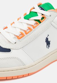 Vit lädersneaker med marinblå detaljer, orange snöröglor och grön tungspets. Har texturerad mocka, framträdande logotyp och gummisula.
