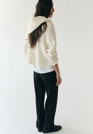 Vrouw met lang donker haar draagt een wit capuchontrui, een wit shirt en een zwarte broek, staand met het gezicht naar een effen witte achtergrond.