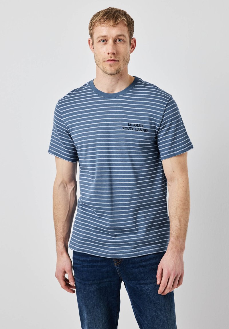 Pruhované modré tričko s krátkými rukávy, které má bílé vodorovné pruhy a malé vyšívané textové logo v černé barvě na hrudi.