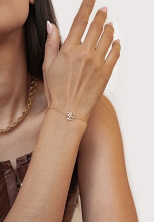 Nahaufnahme eines Handgelenks mit einem zarten goldenen Armband mit einem Baum-Anhänger, Hand nahe am Gesicht erhoben, sichtbares dunkles Haar und braunes Oberteil.