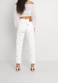 Weiße bestickte Jeans mit floralem Muster, kombiniert mit einem durchsichtigen, schulterfreien, gerafften weißen Oberteil mit puffärmel.