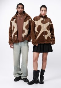 Bruine en beige pluche zip-up jassen met een koeienprint, gecombineerd met lichtblauwe jeans en een zwarte geplooide rok, aangevuld met zwarte kniehoge laarzen.