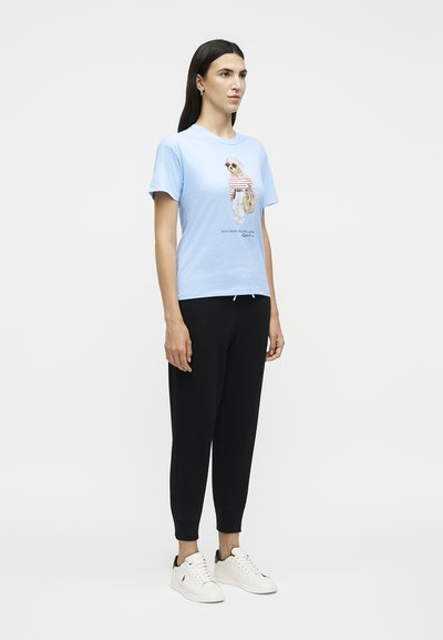 Γυναίκα που φορεί ανοιχτό μπλε μπλουζάκι με σχέδιο αρκούδας, μαύρο κοντό παντελόνι και λευκά αθλητικά παπούτσια, στέκεται μπροστά σε απλό φωτεινό φόντο.