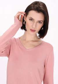Svetlo roza pulover s V-izrezom in dolgimi rokavi; mehak, teksturiran material; manekenka z bob frizuro in zlatimi obročastimi uhani.