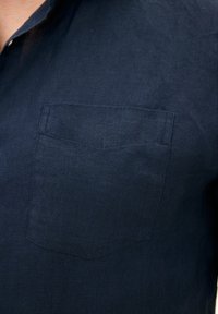 Camicia di lino blu navy con colletto corto, caratterizzata da una tasca a toppa singola sul lato sinistro del petto e un tessuto testurizzato e traspirante.