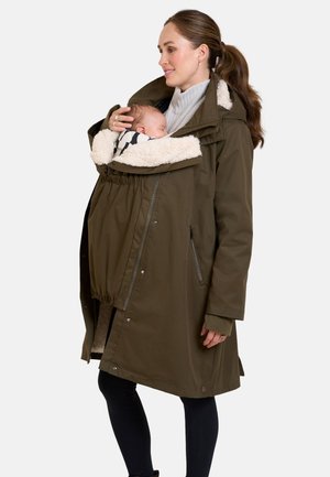Vrouw draagt olijfgroene jas met ingebouwde draagzak en houdt een slapende baby vast die is gewikkeld in een gezellige gevoerde kap.