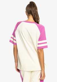 Lekka, kremowa bawełniana koszulka z krótkim rękawem w różowym raglanie i trzema białymi paskami na każdym rękawie. Luźny krój i swobodny design.