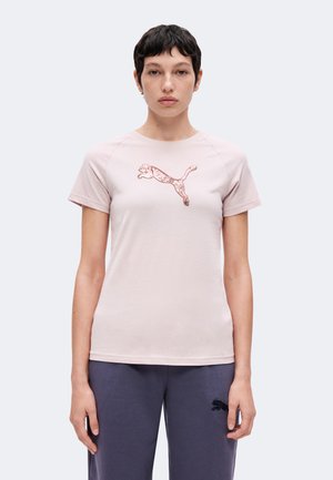 Giovane persona con capelli corti e scuri che indossa una maglietta rosa chiaro a maniche corte con un grande logo Puma e pantaloni scuri con un piccolo logo Puma.