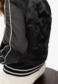 Bomberjack in zwarte satijn met witte gestreepte ribgebreide manchetten en zoom. Heeft een geborduurd draak- en bloemenpatroon op de achterkant.