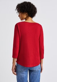 Rotes Langarmoberteil mit weitem Ausschnitt, leicht strukturiertem Stoff und einem abgerundeten Saum. Kombiniert mit hellblauen Jeans.