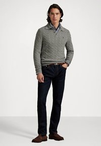 Γκρι πουλόβερ με πλέξη cable και γιακά, σκούρο τζιν, και καφέ μοκασίνια. Το μοντέλο στέκεται με αυτοπεποίθηση με τα χέρια στις τσέπες.