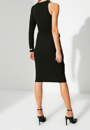 Schwarzes, eng anliegendes Kleid mit einem langen Ärmel und einem schulterfreuen Design; verfügt über einen gerafften Rücken und seitliche Bindebänder. Hohe Absätze.