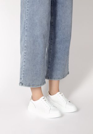Weiße Sneaker mit durchbrochenen Details, einer klobigen weißen Sohle und einem beigen Akzent an der Ferse, kombiniert mit hellblauen Jeans im weiten Schnitt.