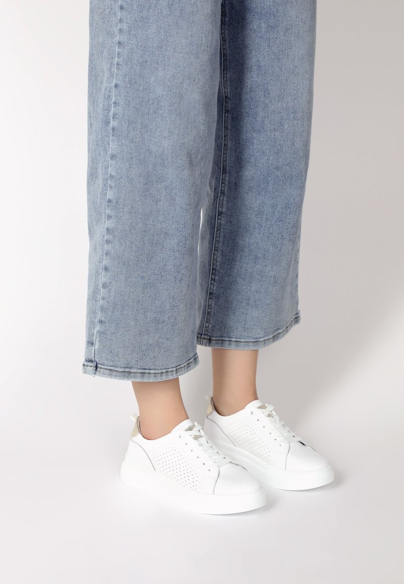 Weiße Sneaker mit durchbrochenen Details, einer klobigen weißen Sohle und einem beigen Akzent an der Ferse, kombiniert mit hellblauen Jeans im weiten Schnitt.