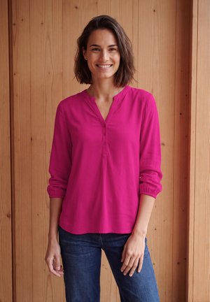 Femme souriante aux cheveux courts et foncés, portant une blouse rose vif à manches longues et un jean bleu foncé, debout contre un mur en bois.