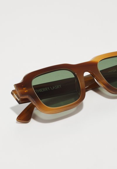 Ochelari de soare transparenți maro cu lentile verzi, design unghiular și ramă groasă. Prezintă inscripția mărcii "THIERRY LARY" pe brațul interior.