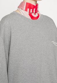 Grå sweatshirt med en sliten nederkant, bärs över en ljusrosa scarf med röda texter och grafiska mönster. Materialen verkar mjuka och avslappnade.