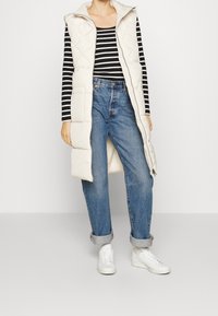 Frau trägt eine weiße gesteppte ärmellose Weste, ein schwarz-weiß gestreiftes langärmeliges Shirt, blaue Jeans mit Umschlag und weiße Turnschuhe.