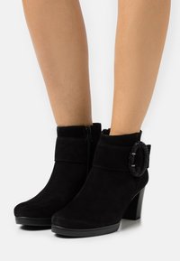 Botines de ante negros con punta redondeada, cremallera lateral, tacón alto y detalle decorativo de cuentas en la correa del tobillo.