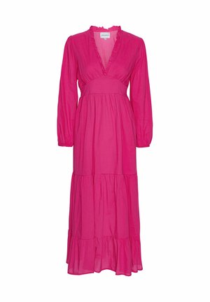 Femme aux cheveux tirés en arrière, portant une robe longue rose vif à manches longues avec un col à volants et une jupe à volants sur fond beige.