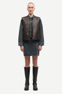 Hnědá kožená vesta přes džínsovou bundu a odpovídající sukni, doplněná vysokými černými botami. Texturované povrchy a kontrastní barvy.