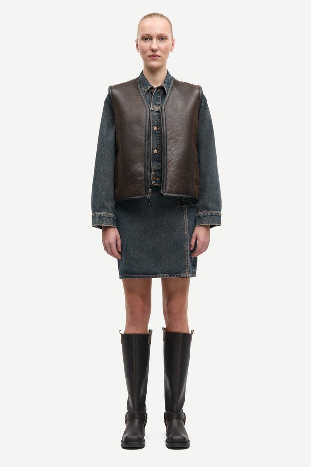 Hnědá kožená vesta přes džínsovou bundu a odpovídající sukni, doplněná vysokými černými botami. Texturované povrchy a kontrastní barvy.
