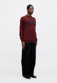 Bordowy sweter z dzianiny z granatowym logo "GCDS" na piersi. Połączony z czarnymi spodniami o szerokich nogawkach i czarnymi butami. Gładkie tło.