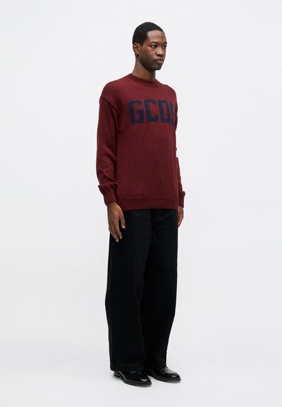 Burgunder strikket genser med et marineblått "GCDS" logo på brystet. Kombinert med svarte vide bukser og svarte sko. Enkel bakgrunn.