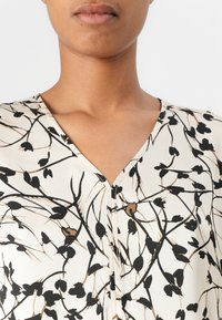 Bluse med blomsterpatter i kremfarge med svarte og gull detaljer. Har V-hals og et glatt, lett stoff. Enkel knappeåpning.
