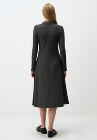 Dunkelgraues, langärmliges Kleid aus weichem, strukturiertem Stoff, mit einem Kragen und in A-Linien-Form, das knapp unter dem Knie endet. Kombiniert mit schwarzen Schuhen.